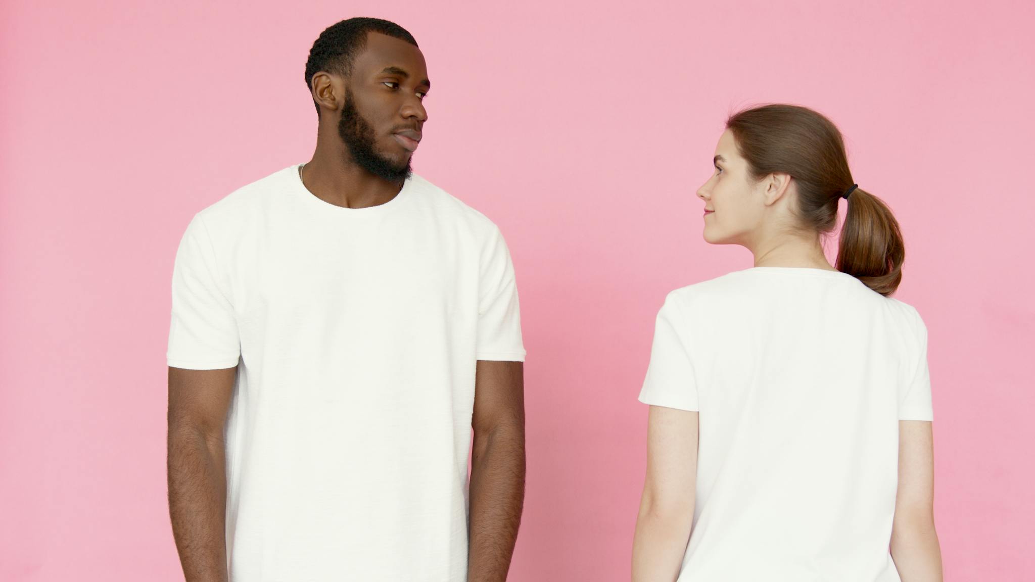 Photo by Artem Podrez A man and woman in white t-shirts stand against a pink backdrop.