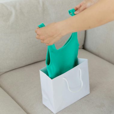 Person unpacking a green tank top from a white shopping bag on a beige couch.