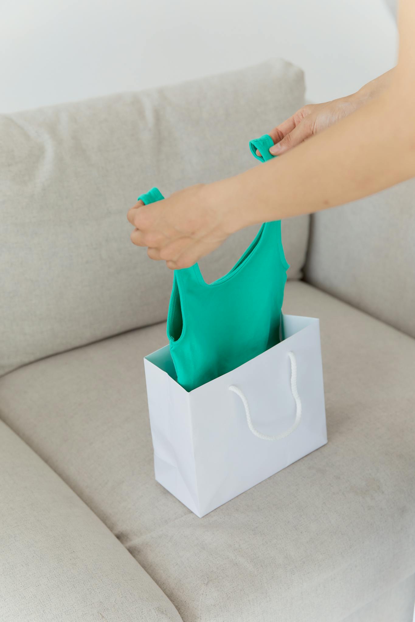 Photo by Darina Belonogova Person unpacking a green tank top from a white shopping bag on a beige couch.