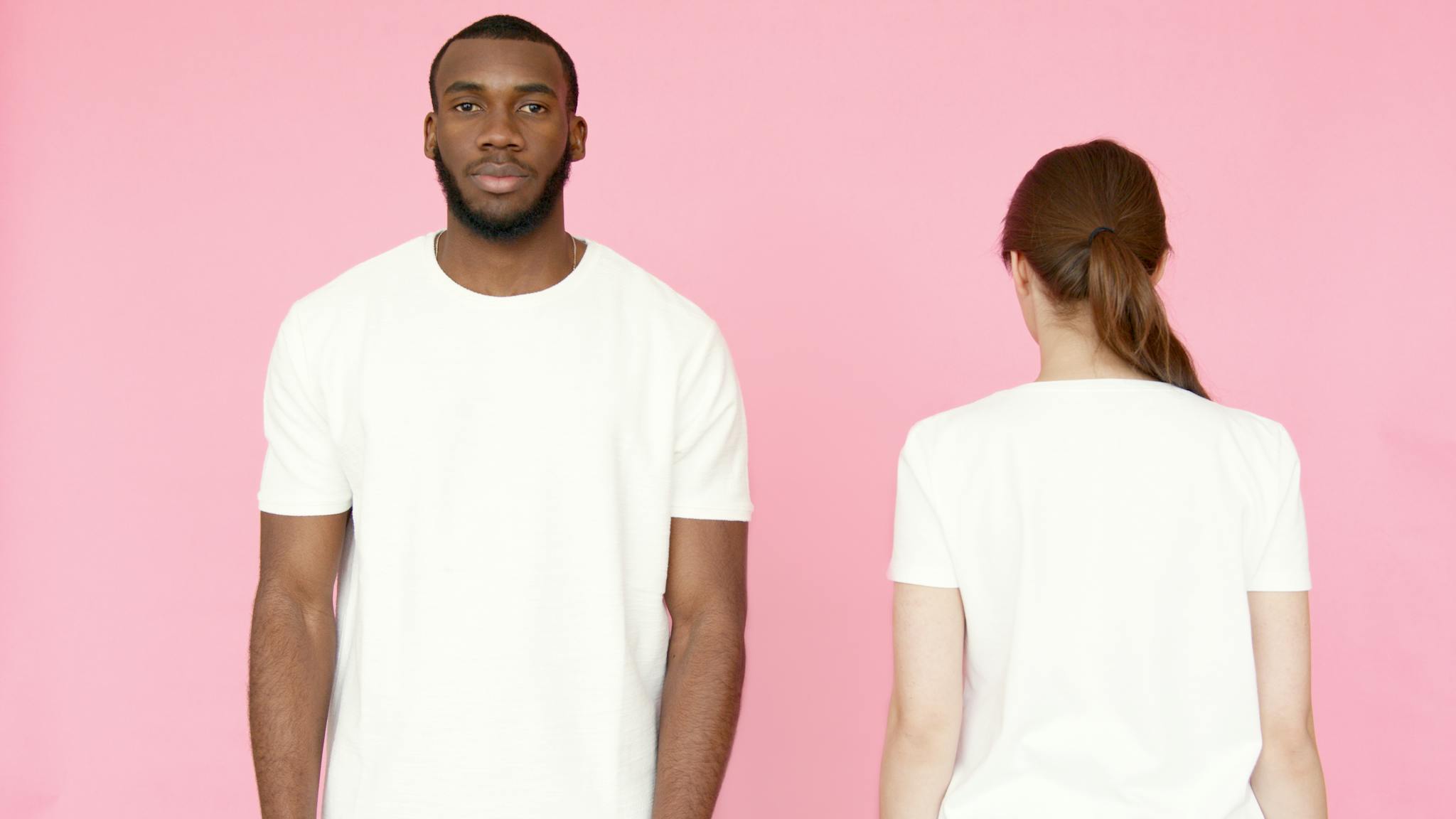 Photo by Artem Podrez Two models showcasing white t-shirts on a pink background, front and back views.